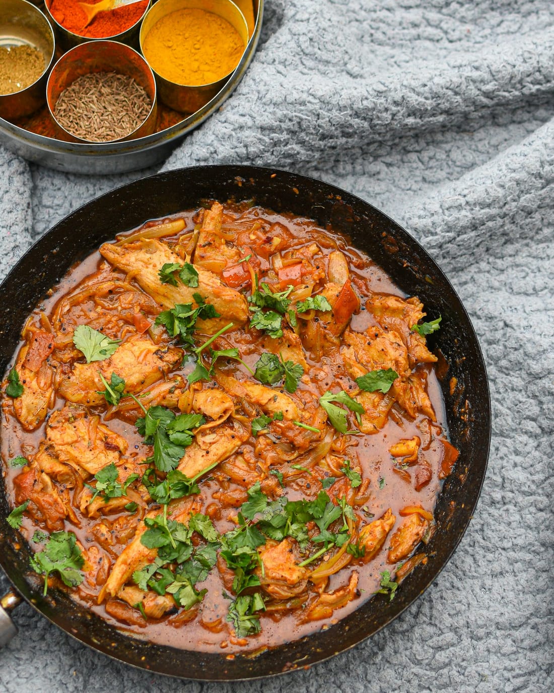 A pan of chicken curry scattered with coriander, seen from above beside a tin of spices.
